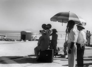 A film crew wearing pith hats, with the director under an umbrella to protect him from the sun